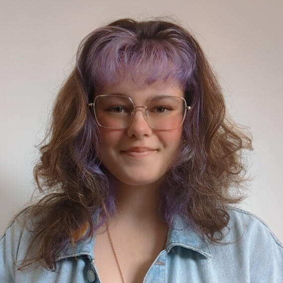 A headshot of Marie Louise Leone, a white woman with brown curly hair dyed purple in the front and wire-rimmed glasses.